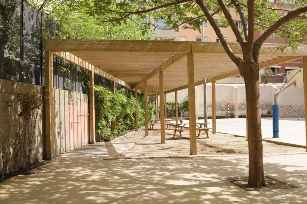 Refugio climático con pérgola de madera y vegetación en un patio escolar